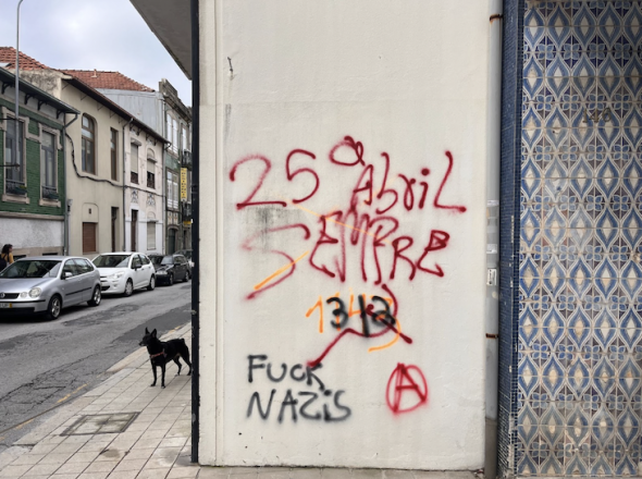 Fotografia de arquivo pessoal, de uma parede no Porto. Há pixos com várias inscrições, em laranja, '1143', por cima, em preto, lê-se '312' e 'Fuck Nazis' e em vermelho '25 de Abril sempre', um símbolo comunista e a letra A num círculo, em referência ao anarquismo