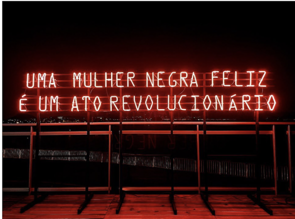 Letreiro em neon obra da artista visual Mônica Ventura com a frase de autoria da escritora e ativista Juliana Borges 'Uma mulher negra feliz é um ato revolucionário'.